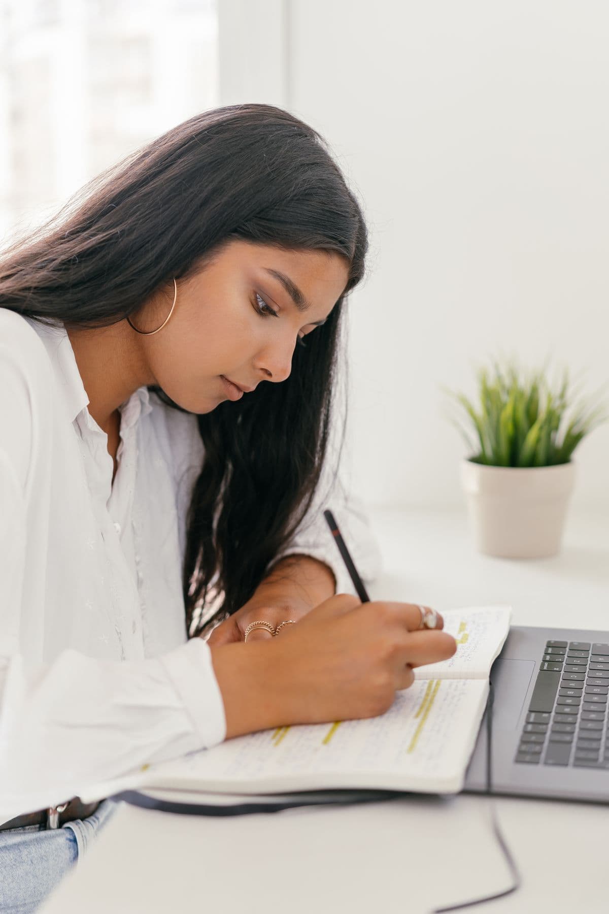 Mulher escrevendo anotações em um caderno ao lado do notebook, organizando as informações dos produtos da loja