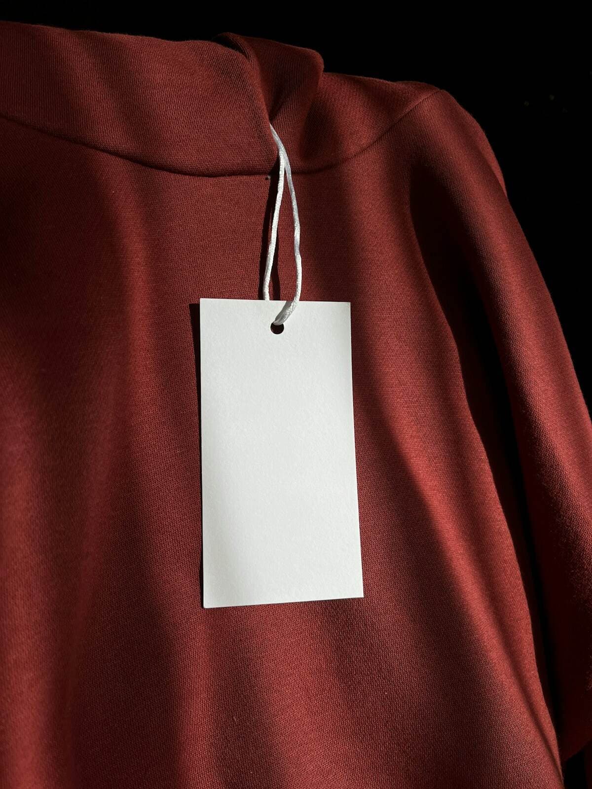 Etiqueta de preço em branco pendurada em uma peça de roupa terracota, representando a dúvida de quanto cobrar por um produto